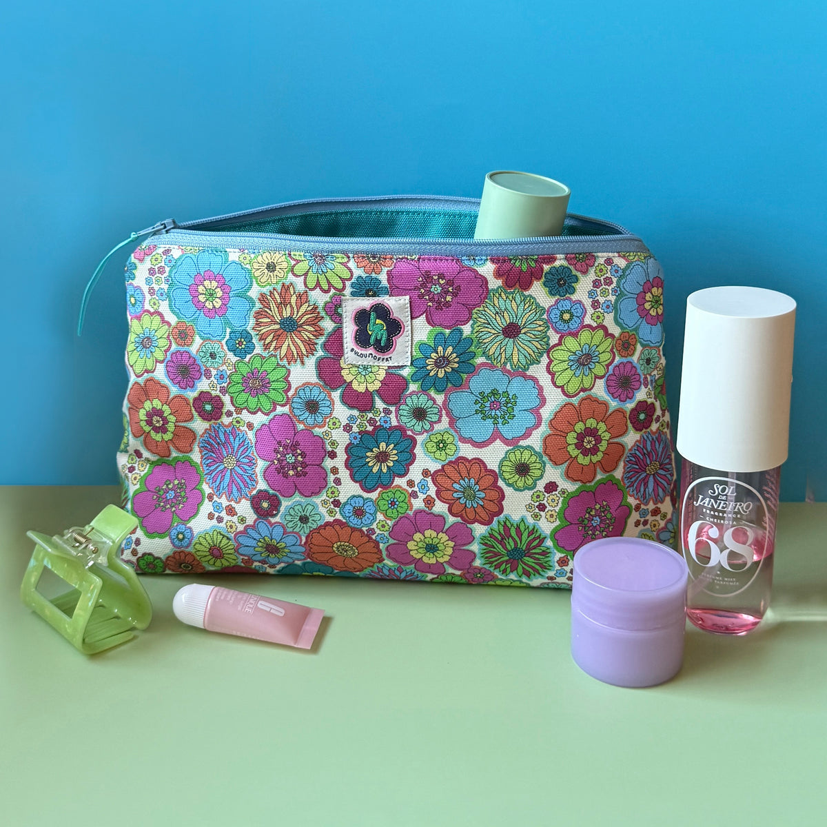 A zippered wash bag/makeup bag with a colourful vintage inspired floral print sits on a light green surface against a bright blue background, surrounded by small beauty accessories including a green hair clip, lip balm tube, purple jar, and nail polish bottle.