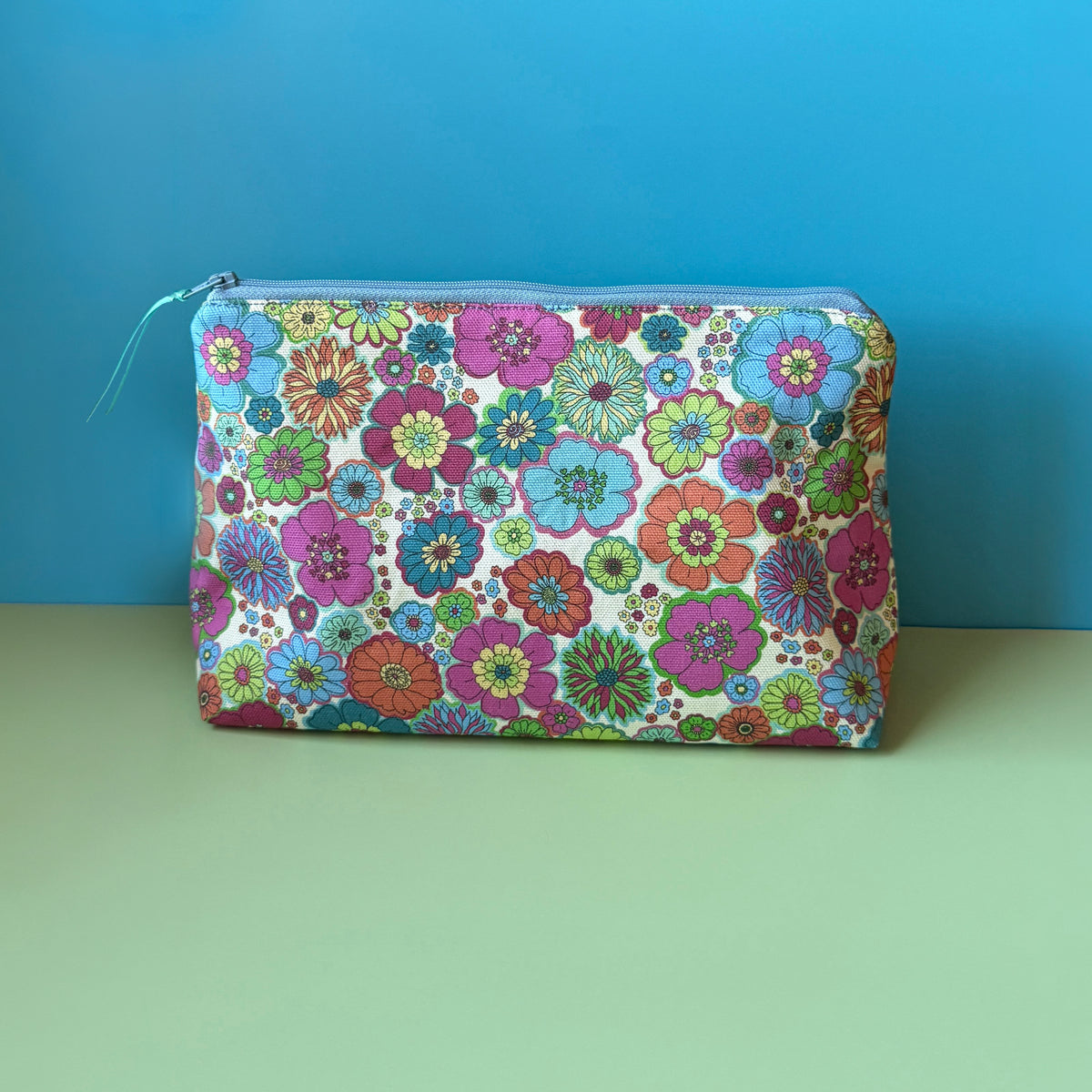 A rectangular zippered pouch with a colourful vintage inspired floral print sits on a light green surface against a bright blue background. The pouch features a dense pattern of pink, blue, green, and orange flowers on a cream base, with a a light blue zipper.
