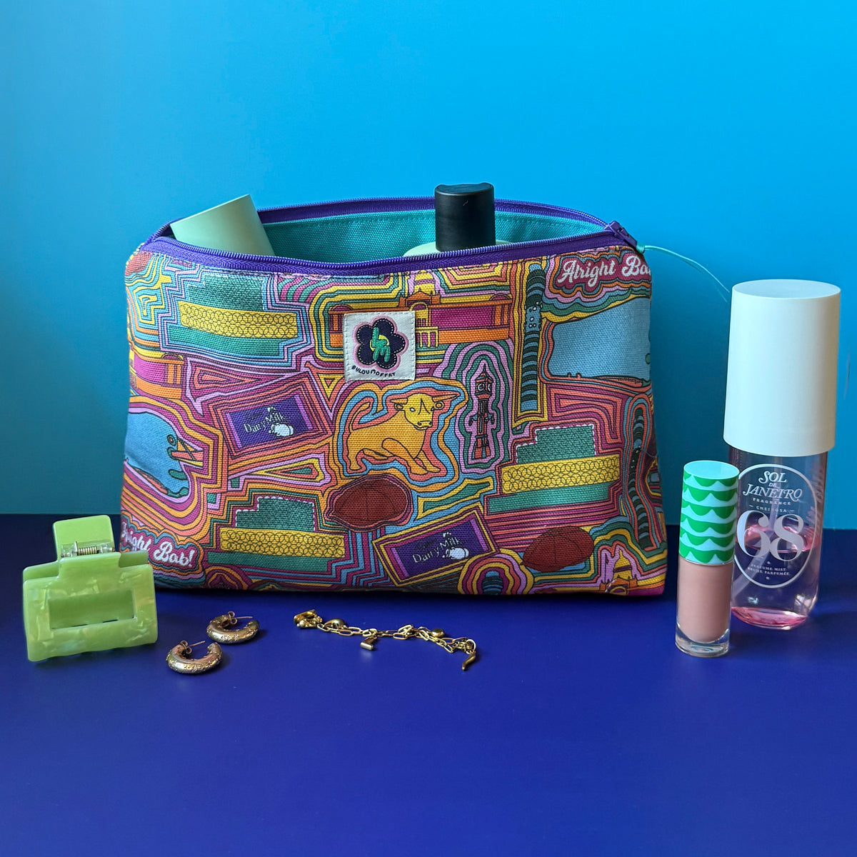 A zippered wash bag/makeup bag with a colourful vintage inspired Birmingham print sits on a royal blue surface against a light blue background, surrounded by small beauty accessories including a green hair clip, lip gloss tube, gold jewellery and sol de janerio pink body spray.