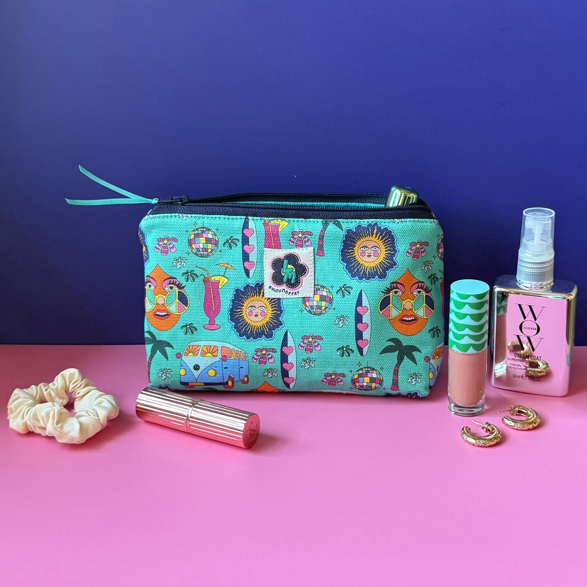 A makeup bag with a hippie design featuring camper vans, surf boards, cocktails, sun, palm tress and disco ball sits open on a pink surface against a royal blue background, surrounded by cosmetic items including a cream scrunchie, gold lipstick, a lipgloss, gold hoop earrings, and mirrored spray bottle.