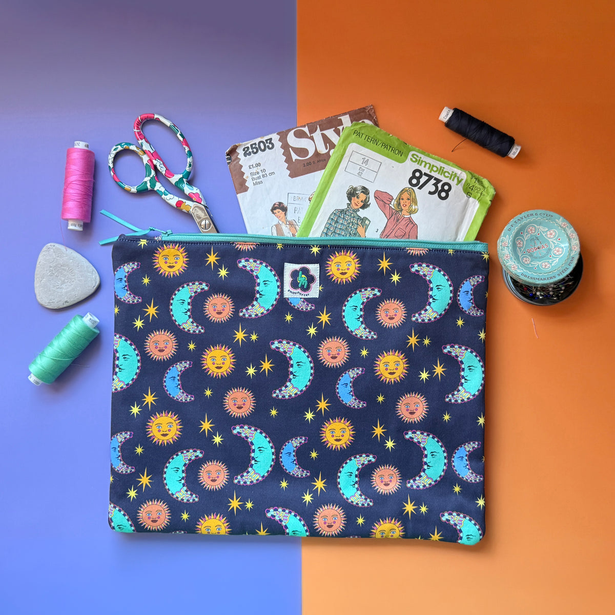 Large navy sun and moon print pouch shown with sewing patterns, scissors, thread and a pin tin. The fabric the pouches is made from features a navy background with teal/blue moons, orange suns and yellow stars.