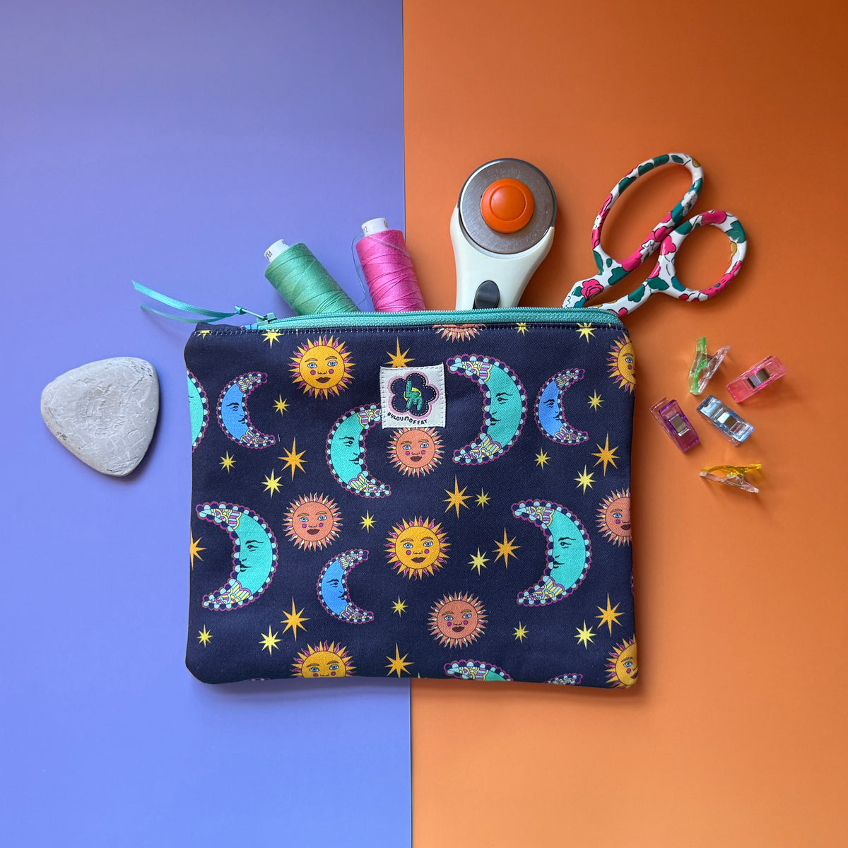 Medium navy sun and moon print pouch shown with thread, scissors, a rotary cutter and sewing clips. The fabric the pouches is made from features a navy background with teal/blue moons, orange suns and yellow stars.