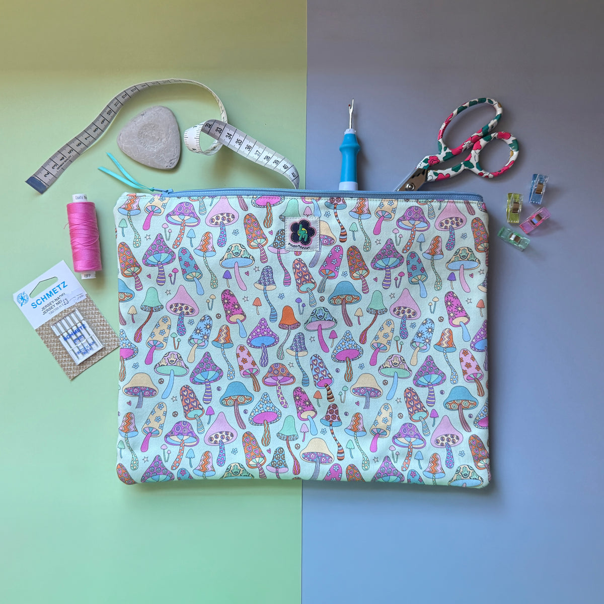 Large mushroom-print zip pouch shown with scissors, thread, a tape measure and sewing accessories. The fabric features colourful patterned mushrooms featuring stars, hearts, flowers hamsa hands.