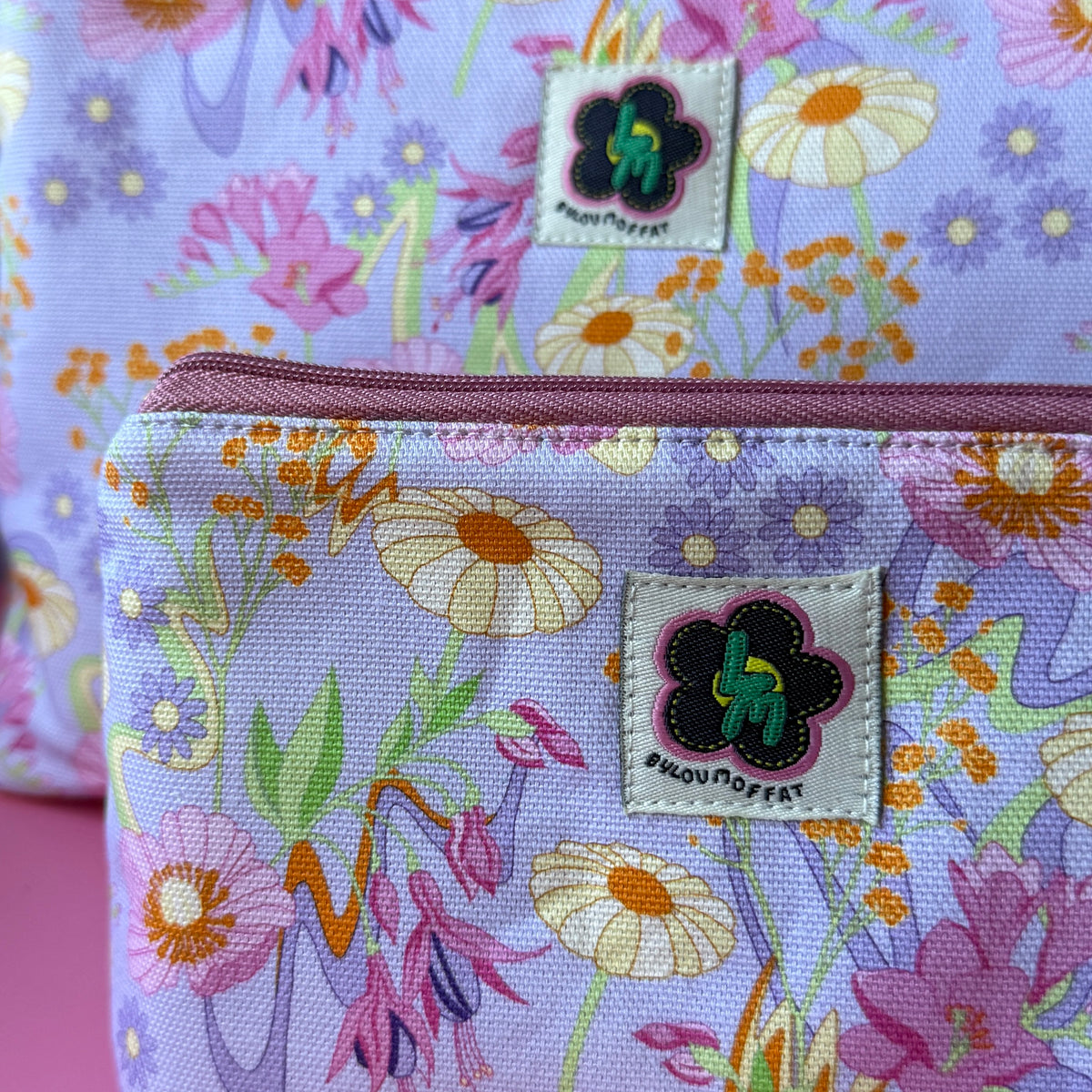 Close-up of a floral zip pouch with two matching bags, featuring a pastel retro flower pattern on a lilac background with a rose zipper and a visible ByLouMoffat brand label.