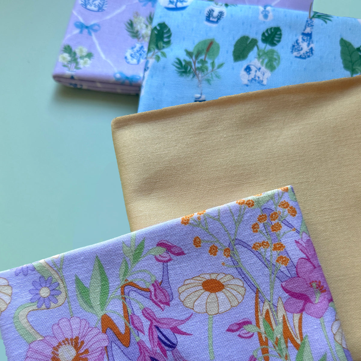 Close-up of retro floral fabric on lilac alongside plain yellow and blue and lilac potted plant fabrics, showing their texture.