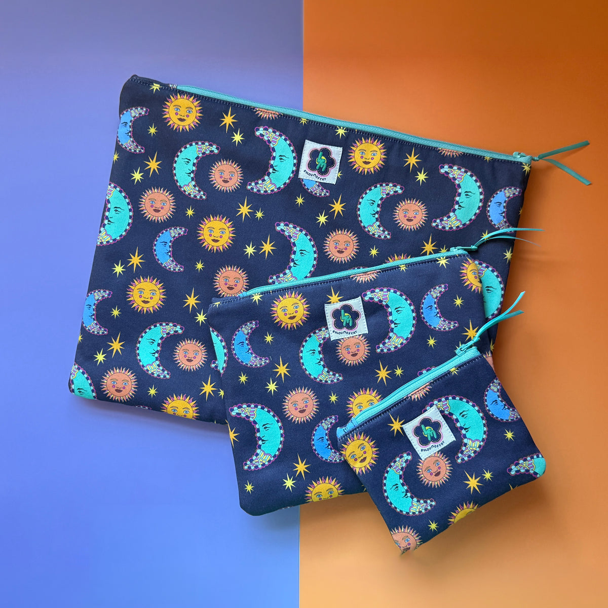 Three navy sun and moon print pouches in small, medium and large sizes, with turquoise zips and teal ribbon. Lying on a 50/50 background of lilac and orange. The fabric the pouches is made from features a navy background with teal/blue moons, orange suns and yellow stars.