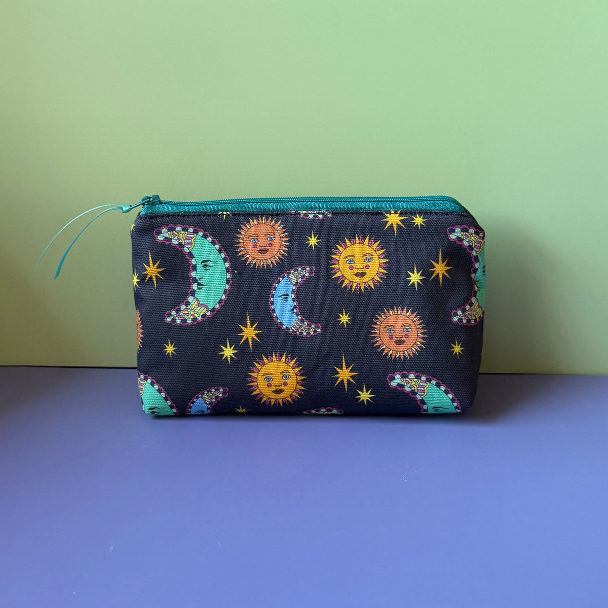 A celestial makeup bag with a navy, orange, blue and teal design that features suns, moons and stars. It has a teal zip with a teal ribbon pull and sits on a lilac surface against a light green background.