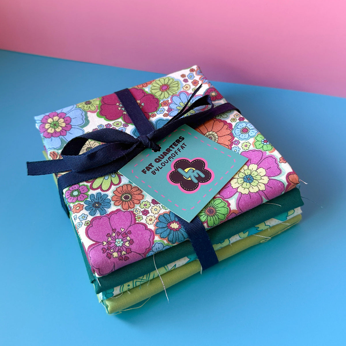  Side view of four stacked fat quarters tied with navy ribbon and a ByLouMoffat tag, with multicoloured floral fabric on top.