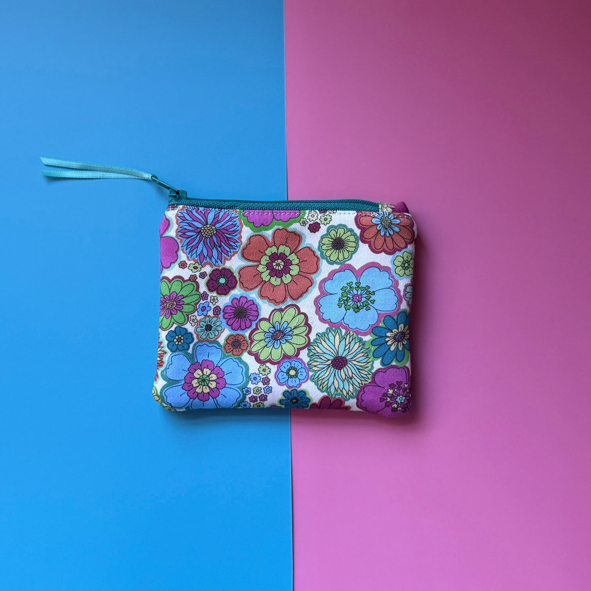 Back of a small zip pouch, showing colourful retro flowers on an off white background with a teal zip.