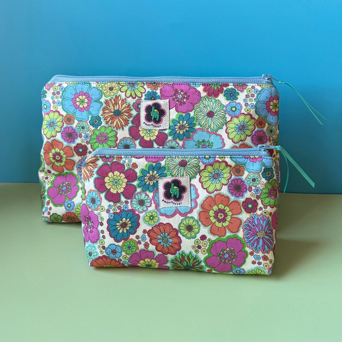 Two fabric zip pouches with a colourful  vintage inspired floral print, ByLouMoffat floral logo labels in the top middle of each bag and blue zip with a teal ribbon pull. Displayed on a light green surface against a blue background.
