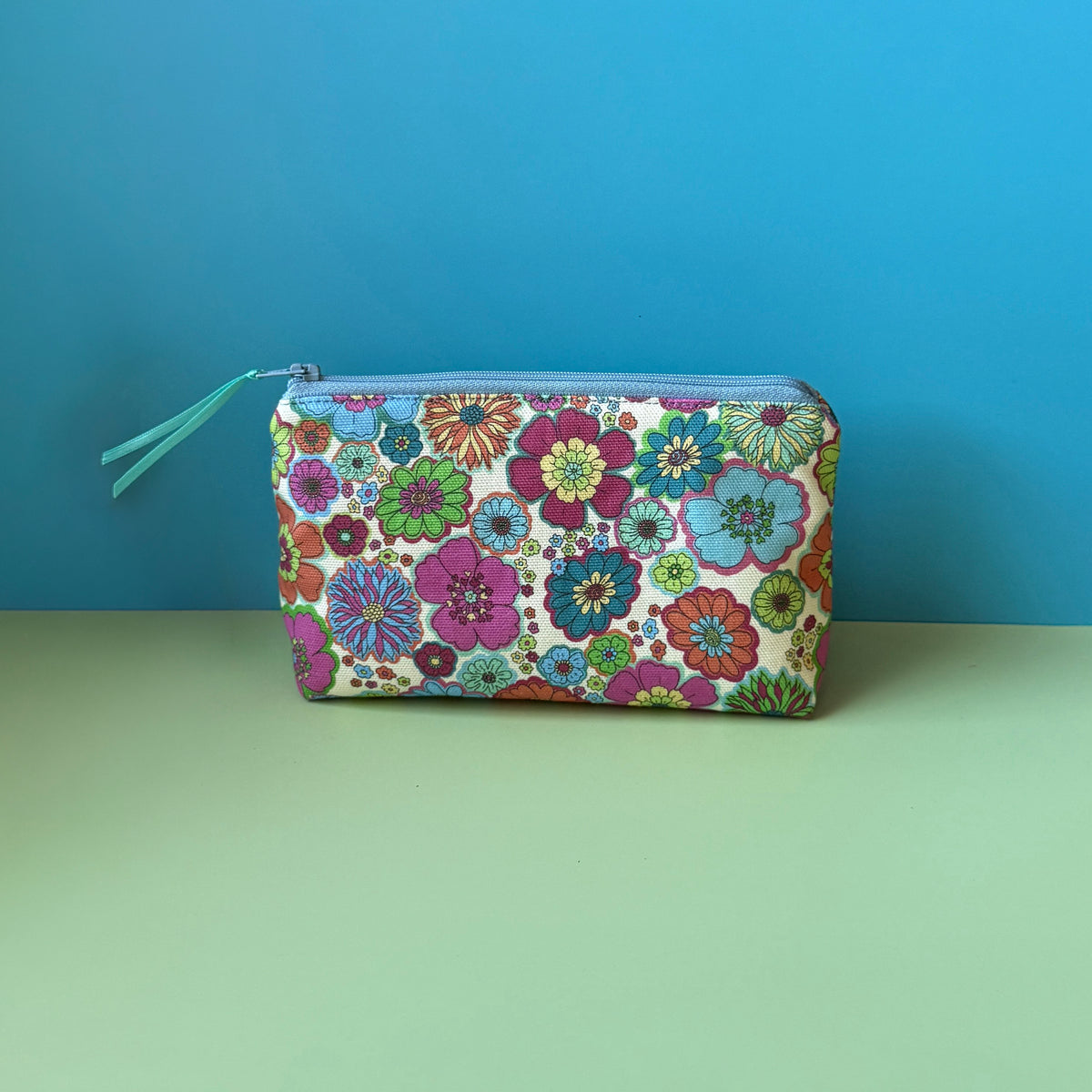 A floral makeup bag with a colourful vintage floral design design and blue zip with teal ribbon sits on a light green surface against a blue background. 