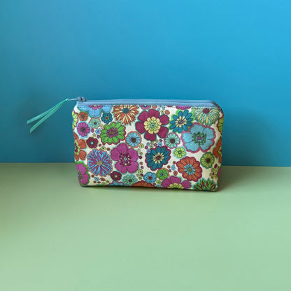 A floral makeup bag with a colourful vintage floral design design and blue zip with teal ribbon sits on a light green surface against a blue background. 