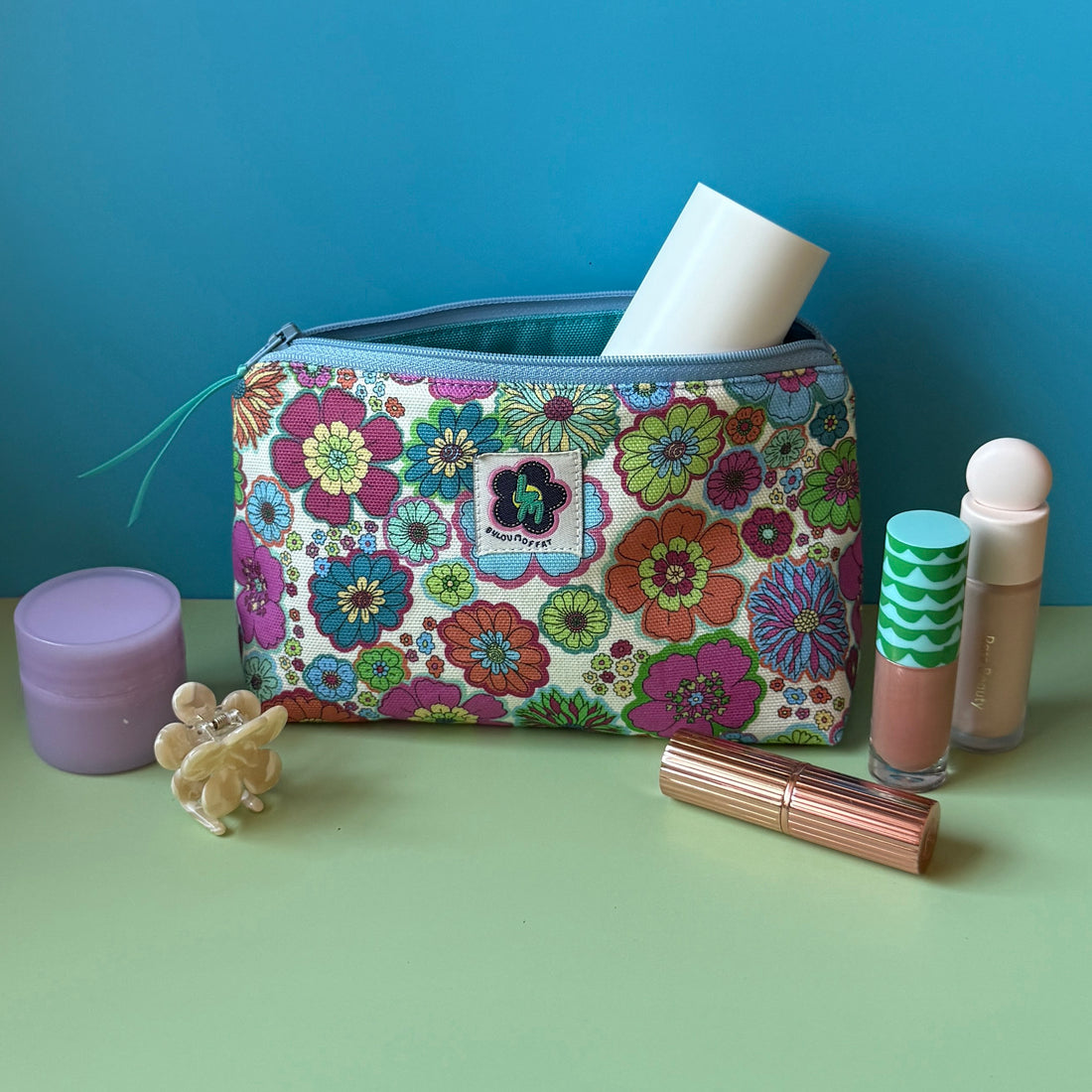 A floral makeup bag with a colourful vintage floral design design sits open on a light green surface against a blue background, surrounded by cosmetic items including a purple moisturiser jar, a small floral hair clip, a lip stick, and a tube of concealer.