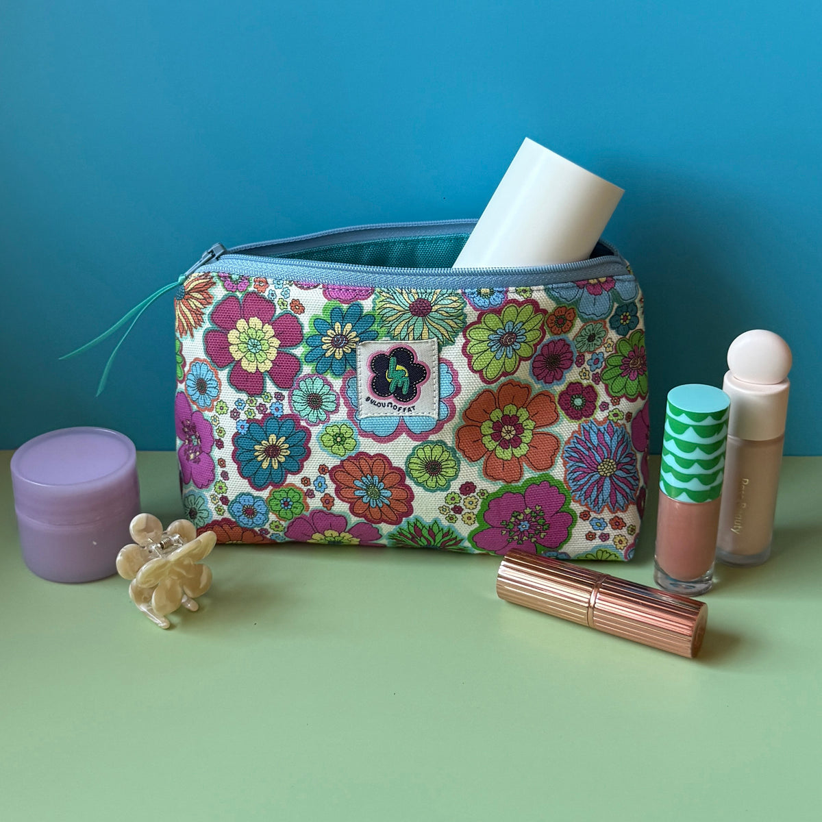 A floral makeup bag with a colourful vintage floral design design sits open on a light green surface against a blue background, surrounded by cosmetic items including a purple moisturiser jar, a small floral hair clip, a lip stick, and a tube of concealer.