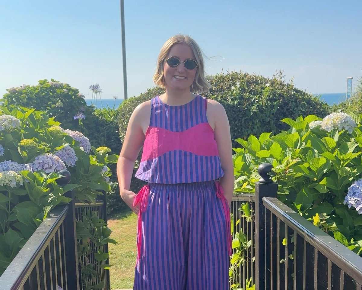 A woman wearing a purple and pink striped sleeveless jumpsuit walks down a wooden boardwalk outdoors, surrounded by green hydrangea bushes under a clear blue sky.
