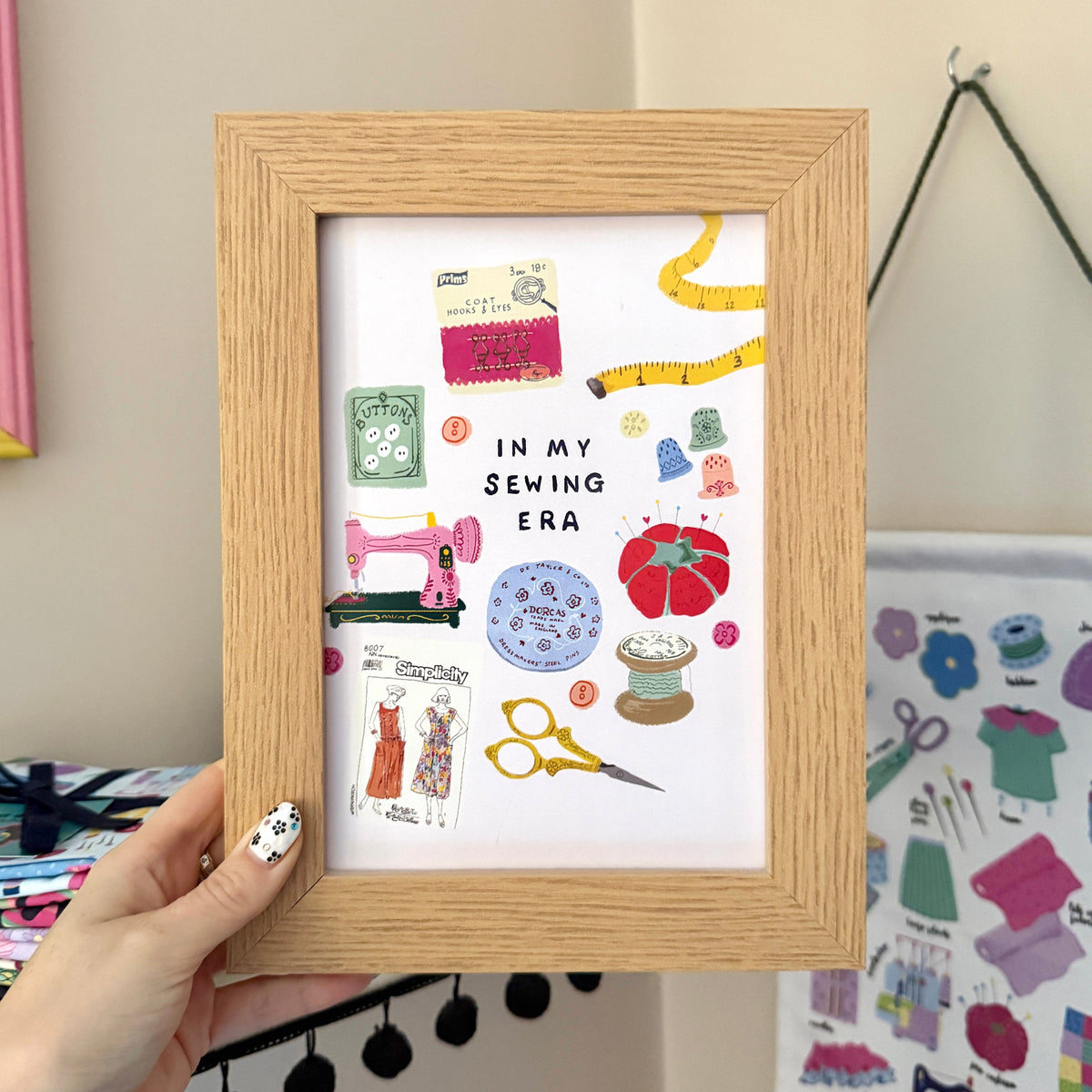 Hand holding an ‘In My Sewing Era’ print in a wooden frame, featuring a pink sewing machine and colourful vintage inspired sewing notions.