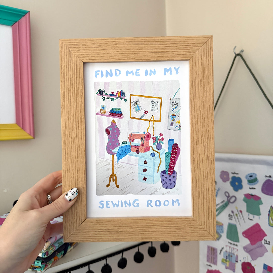 Hand holding a ‘Find Me In My Sewing Room’ print in a wooden frame, featuring a colourful illustrated sewing space with a mannequin, fabric rolls and more.