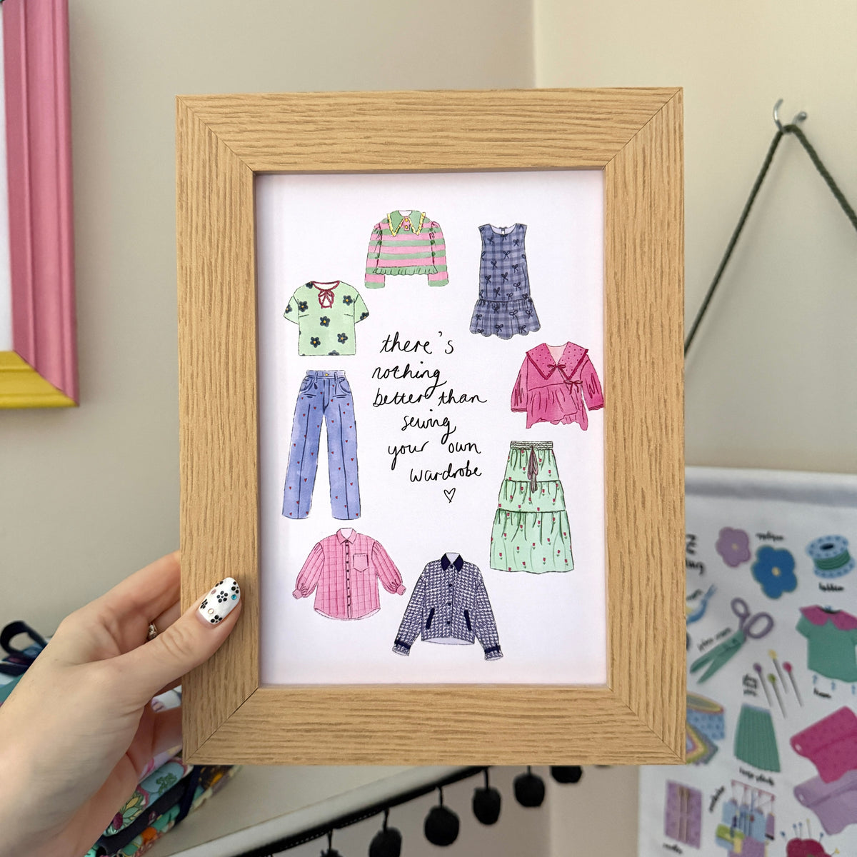 Hand holding a wooden-framed print with the words ‘There’s nothing better than sewing your own wardrobe’, surrounded by illustrated clothes in pink, green and purple.