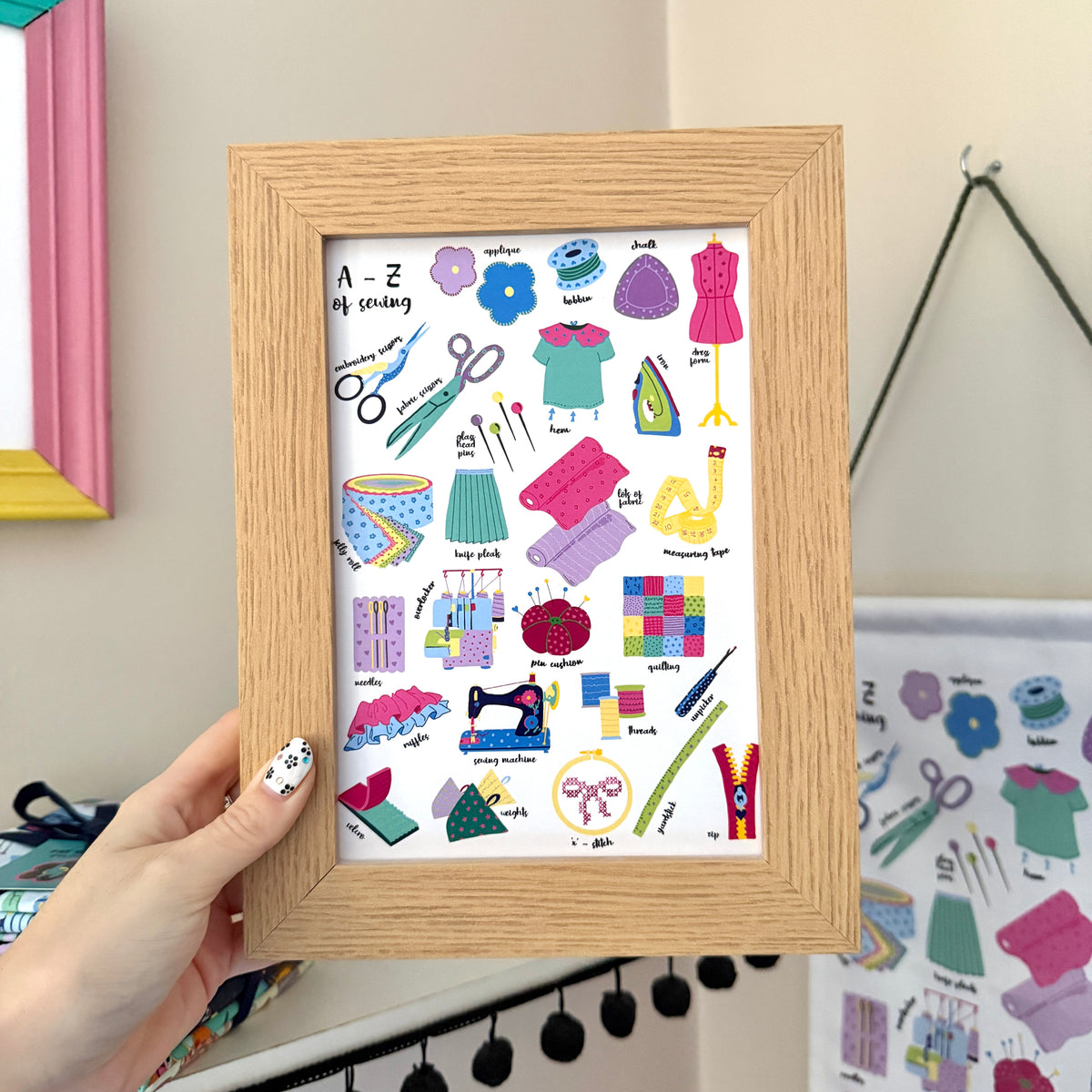 Hand holding an A–Z of Sewing print in a wooden frame, featuring colourful illustrations of sewing tools, fabric and techniques.