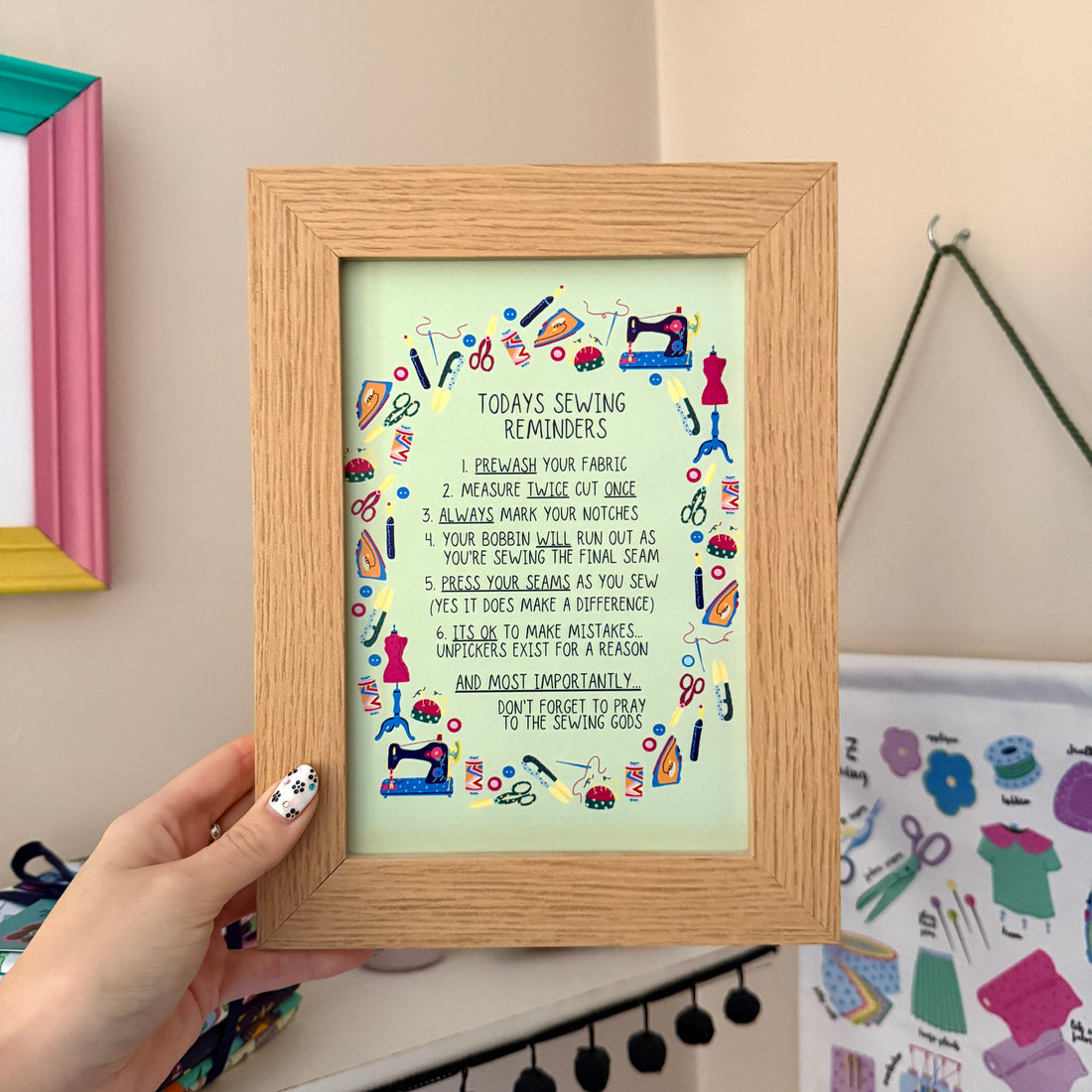Hand holding a Sewing Reminders print in a wooden frame, with sewing tips surrounded by colourful illustrations of sewing tools.