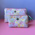
Two fabric zip pouches with a pastel retro inspired floral print, ByLouMoffat floral logo labels in the top middle of each bag and rose coloured zip with a teal ribbon pull. Displayed on a pink surface against a lilac background.
