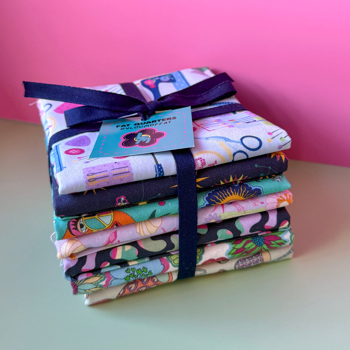 Seven printed fat quarters stacked and tied with a navy ribbon and turquoise ByLouMoffat tag, with the sewing-themed fabric on top.