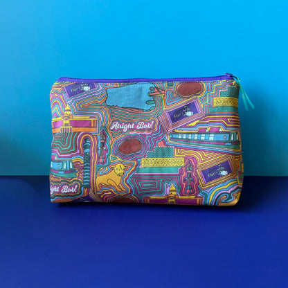 A rectangular zippered pouch with a colourful vintage inspired Birmingham print sits on a royal blue surface against a light blue background. The pouch features a dense pattern of Birmingham icons including, a canal boat, Peahy Blinder hat, the library, BT tower, Selfridges building, Cadburys bar, the Bull and the slogan &