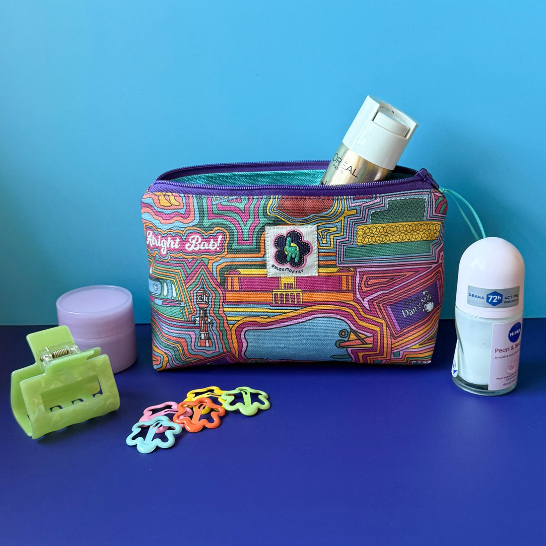 A wash bag/makeup bag with a colourful vintage Birmingham design sits open on a royal blue surface against a light blue background, surrounded by cosmetic items including a purple moisturiser jar, a series of small floral hair clips, a mini deodorant and a green claw clip. 