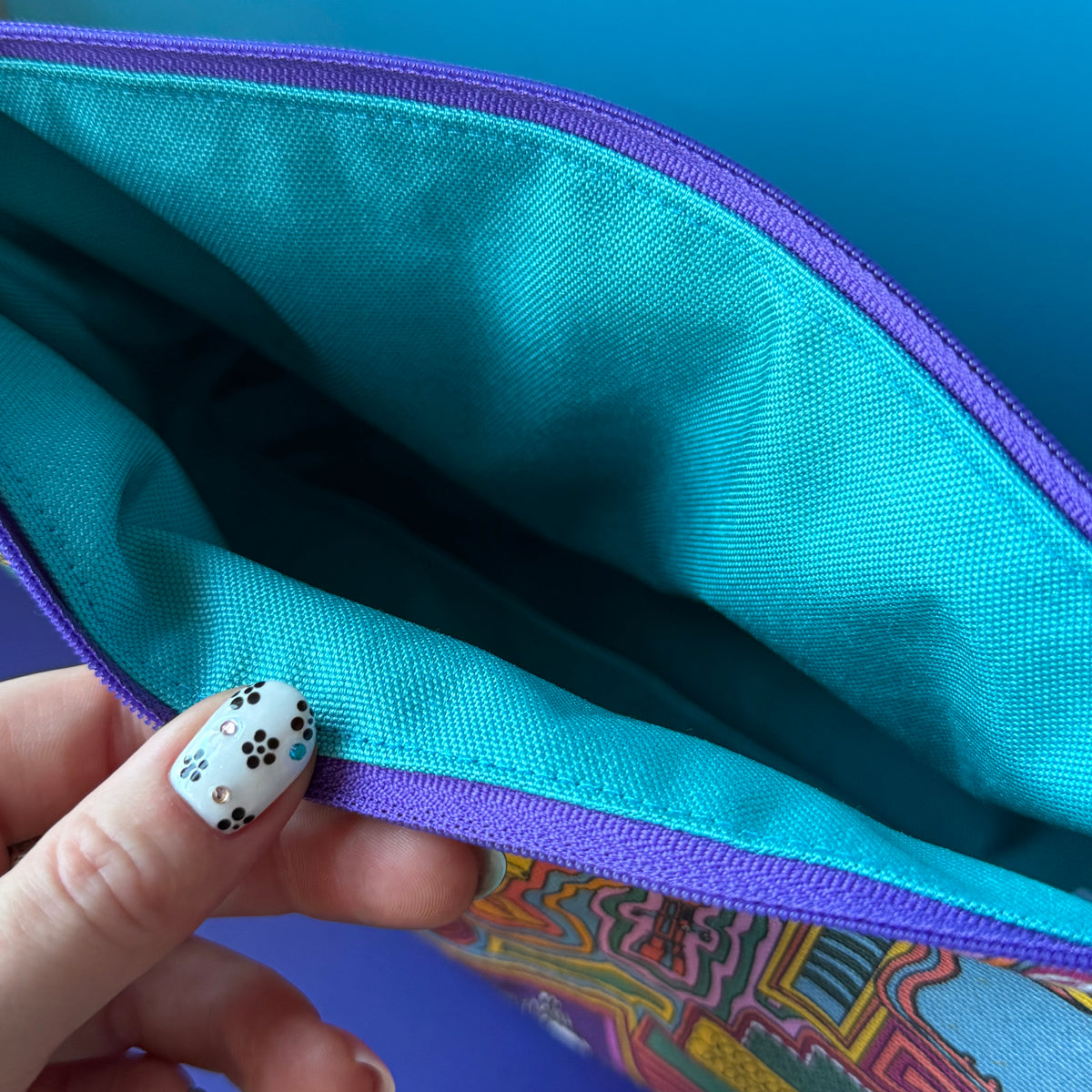 Close-up of a zippered pouch opened by a hand with black-and-white floral nail art, showing a turquoise water repellant interior and a multicolor exterior.