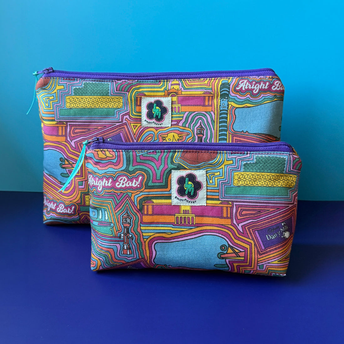 Two fabric zip pouches with a colourful  retro inspired Birmingham based print, ByLouMoffat floral logo labels in the top middle of each bag and purple zip with a teal ribbon pull. Displayed on a royal blue surface against a light blue background.