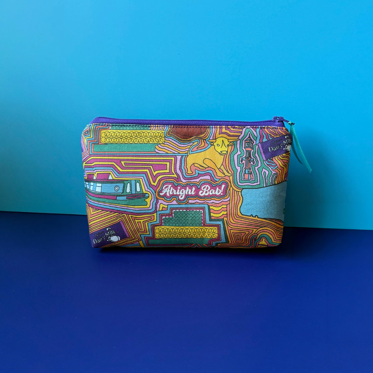 A wash bag/makeup bag with a colourful vintage Birmingham inspired design and purple zip with teal ribbon sits on royal blue green surface against a light blue background. 