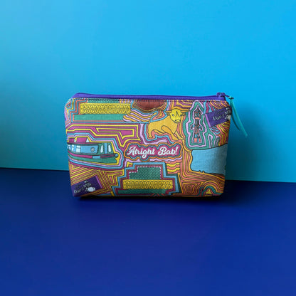 A wash bag/makeup bag with a colourful vintage Birmingham inspired design and purple zip with teal ribbon sits on royal blue green surface against a light blue background. 