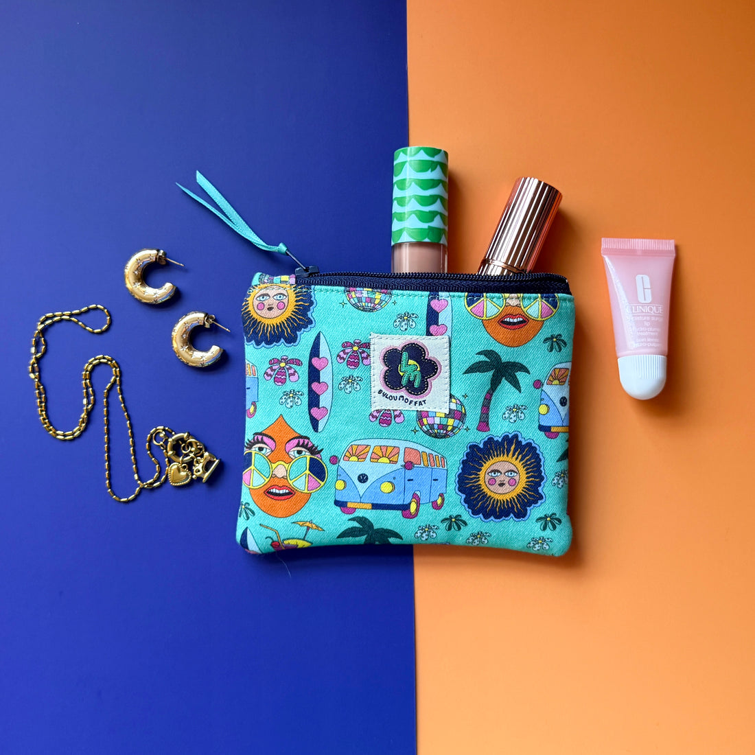 Small turquoise Hippie Pouch with campervan, disco ball, palm tree and surfboard and peace-sign print, shown with cosmetics and gold jewellery.