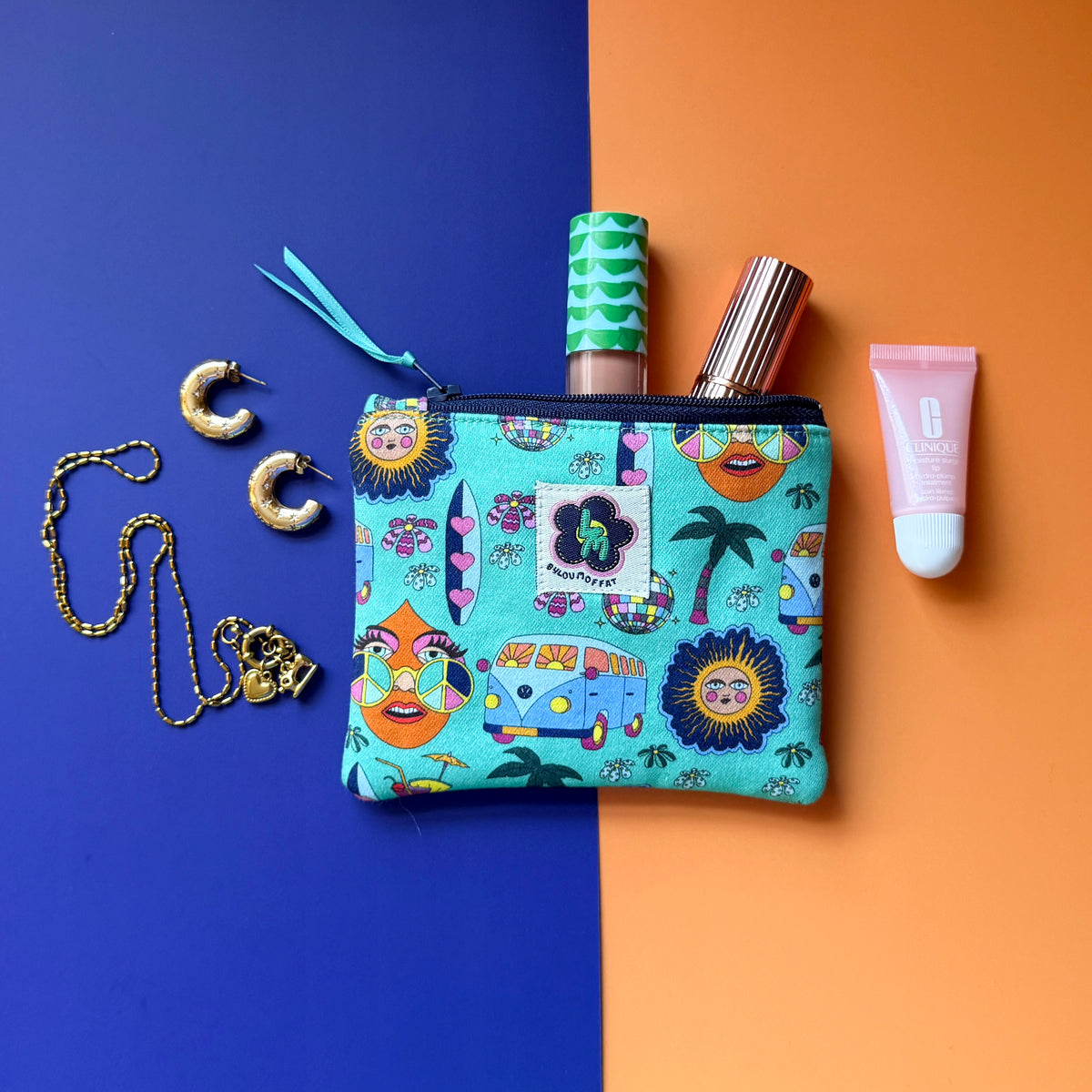 Small turquoise Hippie Pouch with campervan, disco ball, palm tree and surfboard and peace-sign print, shown with cosmetics and gold jewellery.
