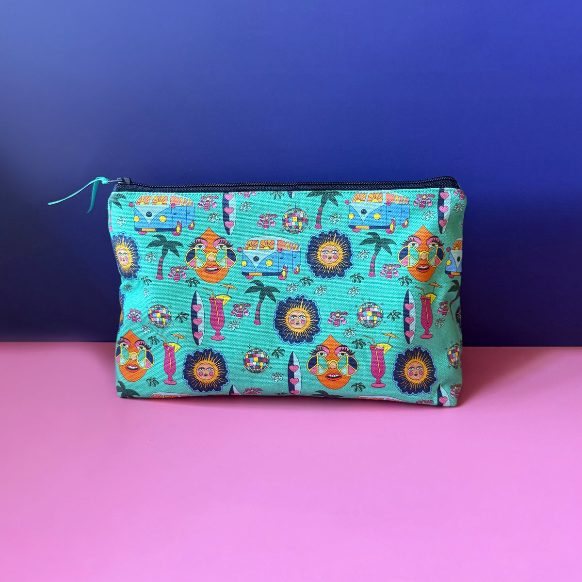 A rectangular zippered pouch with a colourful vintage inspired hippie print with teal background sits on a pink surface against a royal blue backdrop. The pouch features a dense pattern of camper vans, surf boards, cocktails, sun, palm tress and disco ball. on a teal base, with a navy zipper.