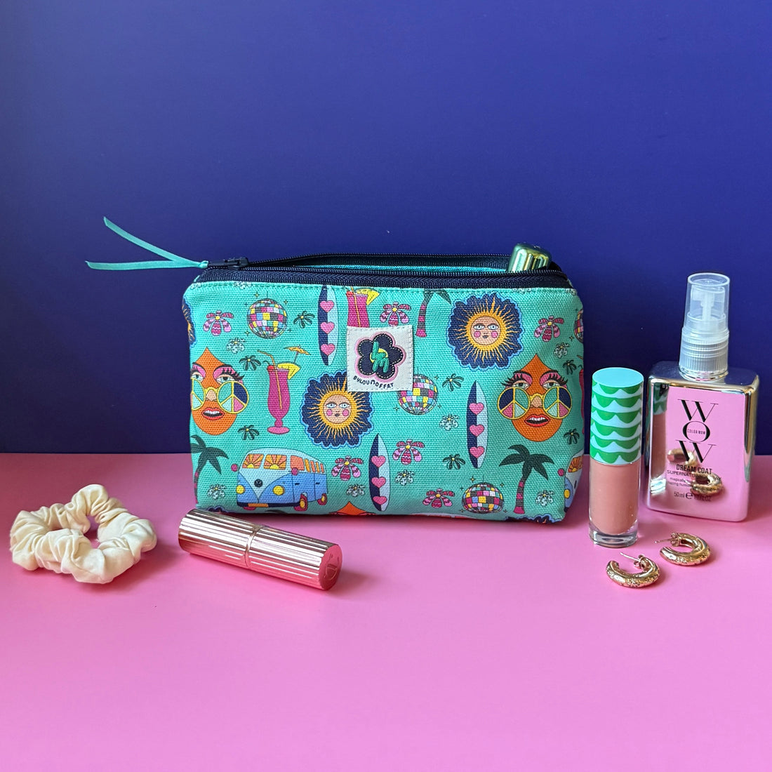 A makeup bag with a hippie design featuring camper vans, surf boards, cocktails, sun, palm tress and disco ball sits open on a pink surface against a royal blue background, surrounded by cosmetic items including a cream scrunchie, gold lipstick, a lipgloss, gold hoop earrings, and mirrored spray bottle.