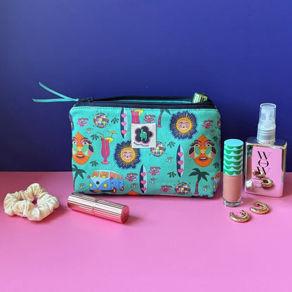 A makeup bag with a hippie design featuring camper vans, surf boards, cocktails, sun, palm tress and disco ball sits open on a pink surface against a royal blue background, surrounded by cosmetic items including a cream scrunchie, gold lipstick, a lipgloss, gold hoop earrings, and mirrored spray bottle.