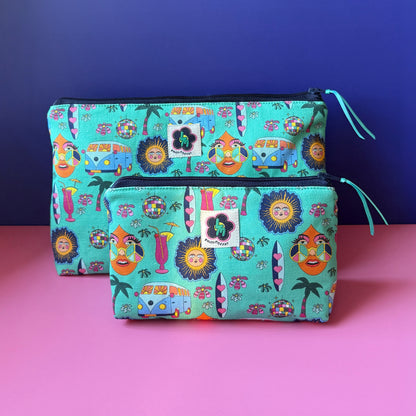 Two fabric zip pouches with a colourful  hippie inspired print, ByLouMoffat floral logo labels in the top middle of each bag and navy zip with a teal ribbon pull. Displayed on a pink surface against a royal blue background.
