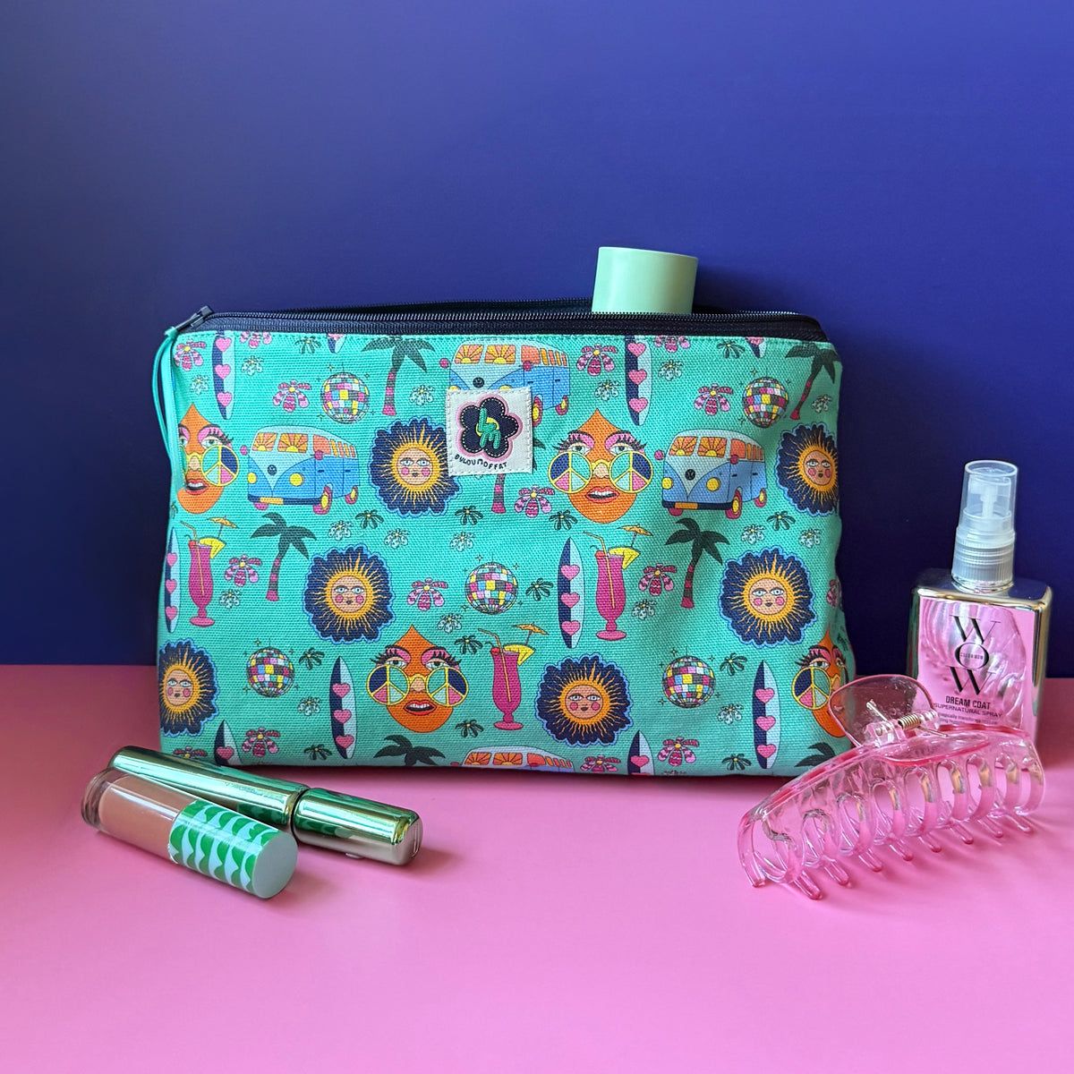 A  zippered wash bag/makeup bag with a colourful hippie inspired print sits on a pink surface against a royal blue background, surrounded by small beauty accessories including a pink hair clip, lip gloss tube, green mascara, and mirrored spray bottle. 