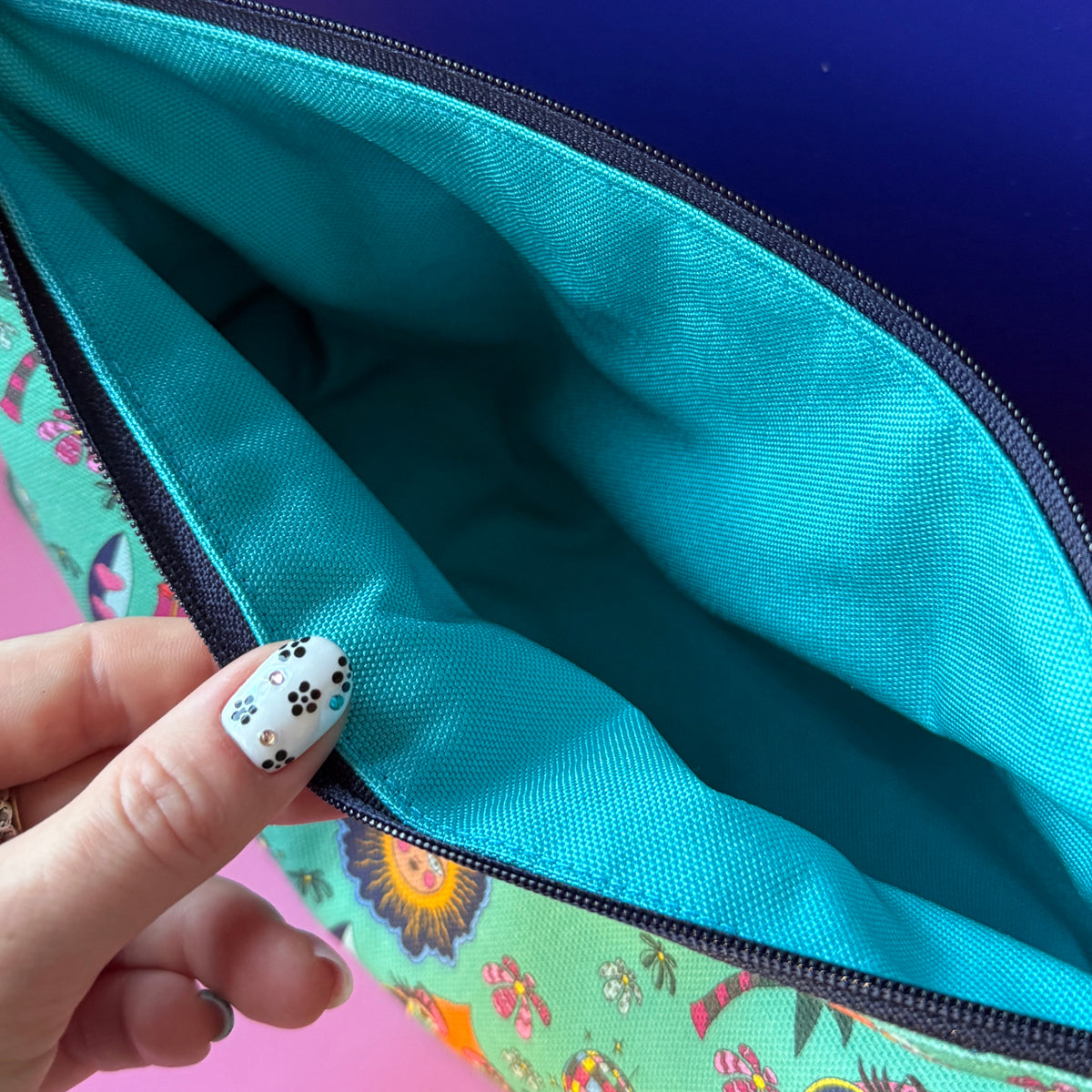 Close-up of a zippered pouch opened by a hand with black-and-white floral nail art, showing a turquoise water repellant interior and a multicolor hippie exterior.