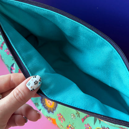 Close-up of a zippered pouch opened by a hand with black-and-white floral nail art, showing a turquoise water repellant interior and a multicolor hippie exterior.