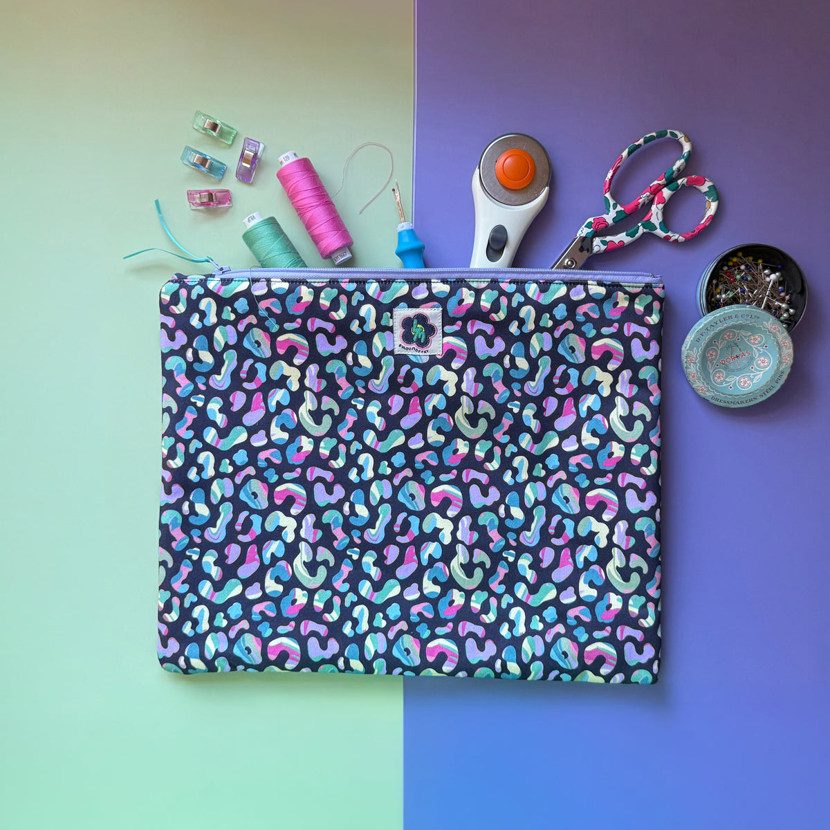 Large leopard print zip pouch shown with scissors, thread, a rotary cutter and sewing accessories. Fabric features colourful leopard print spots on a dark navy background. 