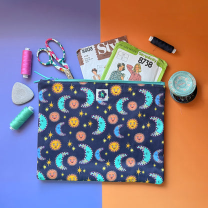 Large navy sun and moon print pouch shown with sewing patterns, scissors, thread and a pin tin. The fabric the pouches is made from features a navy background with teal/blue moons, orange suns and yellow stars. 