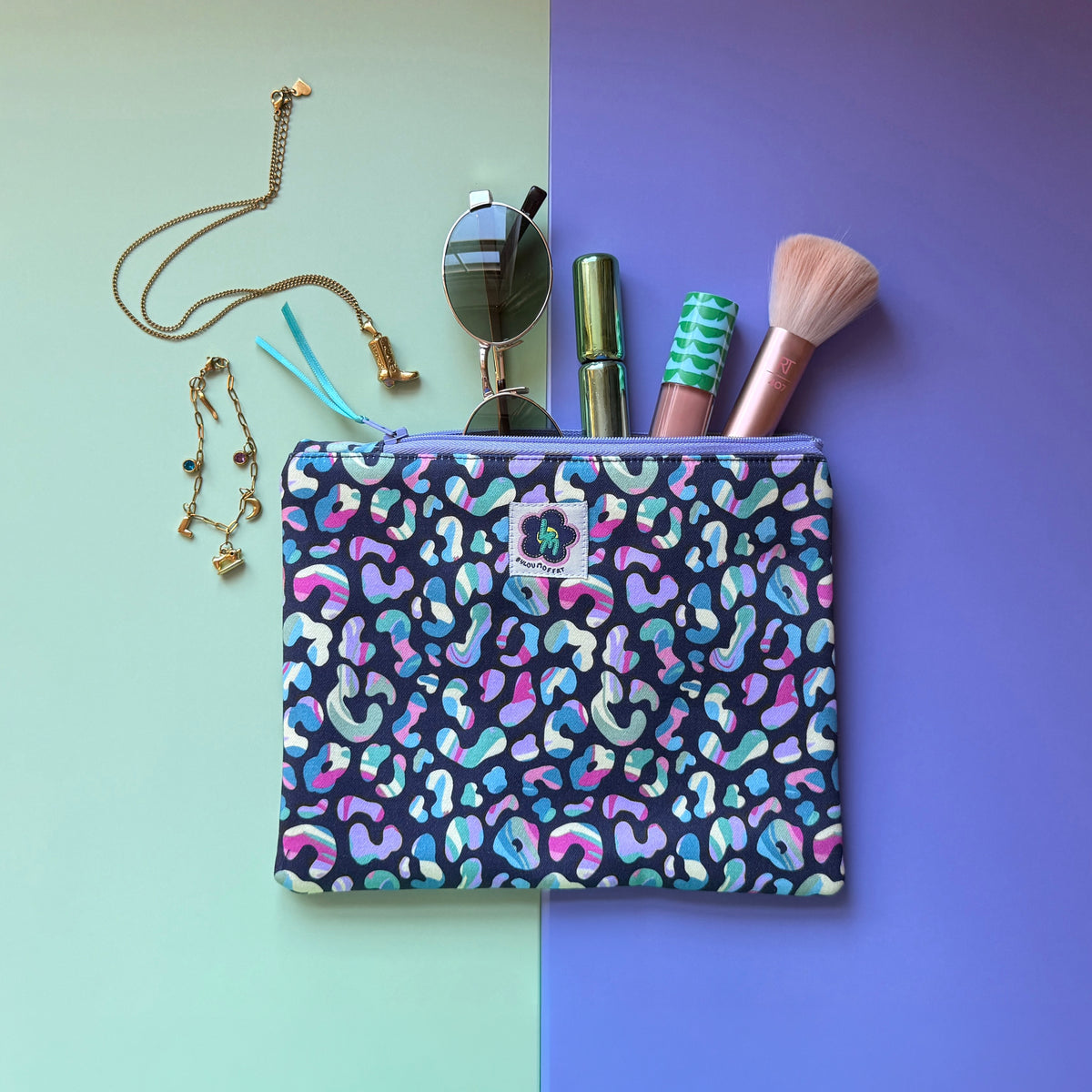 Medium leopard print zip pouch featuring colourful leopard spots on a navy background. Photographed with sunglasses, make-up, a brush and jewellery.