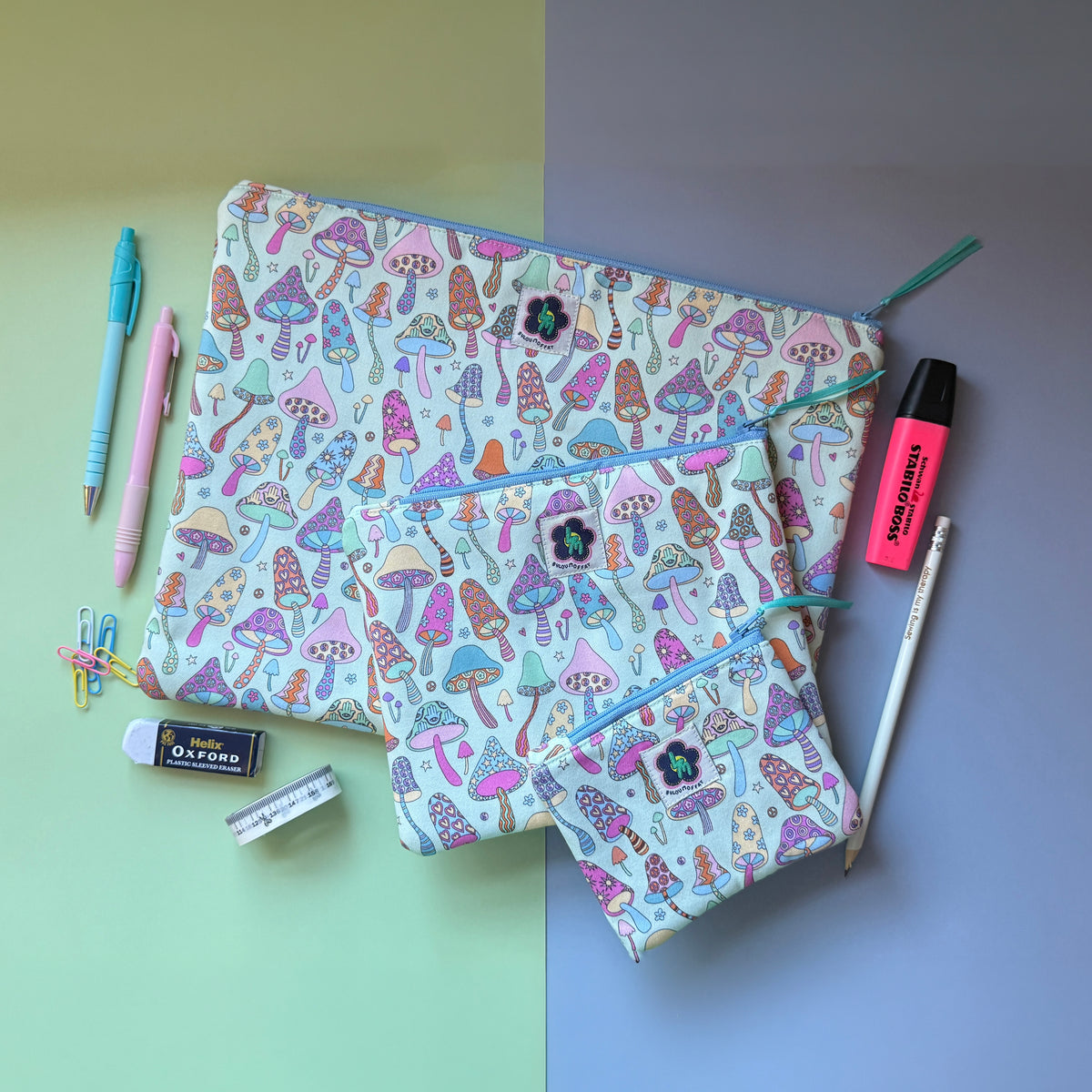 Three matching mushroom-print zip pouches arranged with pens, a highlighter, pencil and other stationery. The fabric features colourful patterned mushrooms featuring stars, hearts, flowers hamsa hands. 