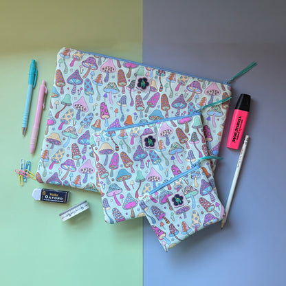 Three matching mushroom-print zip pouches arranged with pens, a highlighter, pencil and other stationery. The fabric features colourful patterned mushrooms featuring stars, hearts, flowers hamsa hands. 