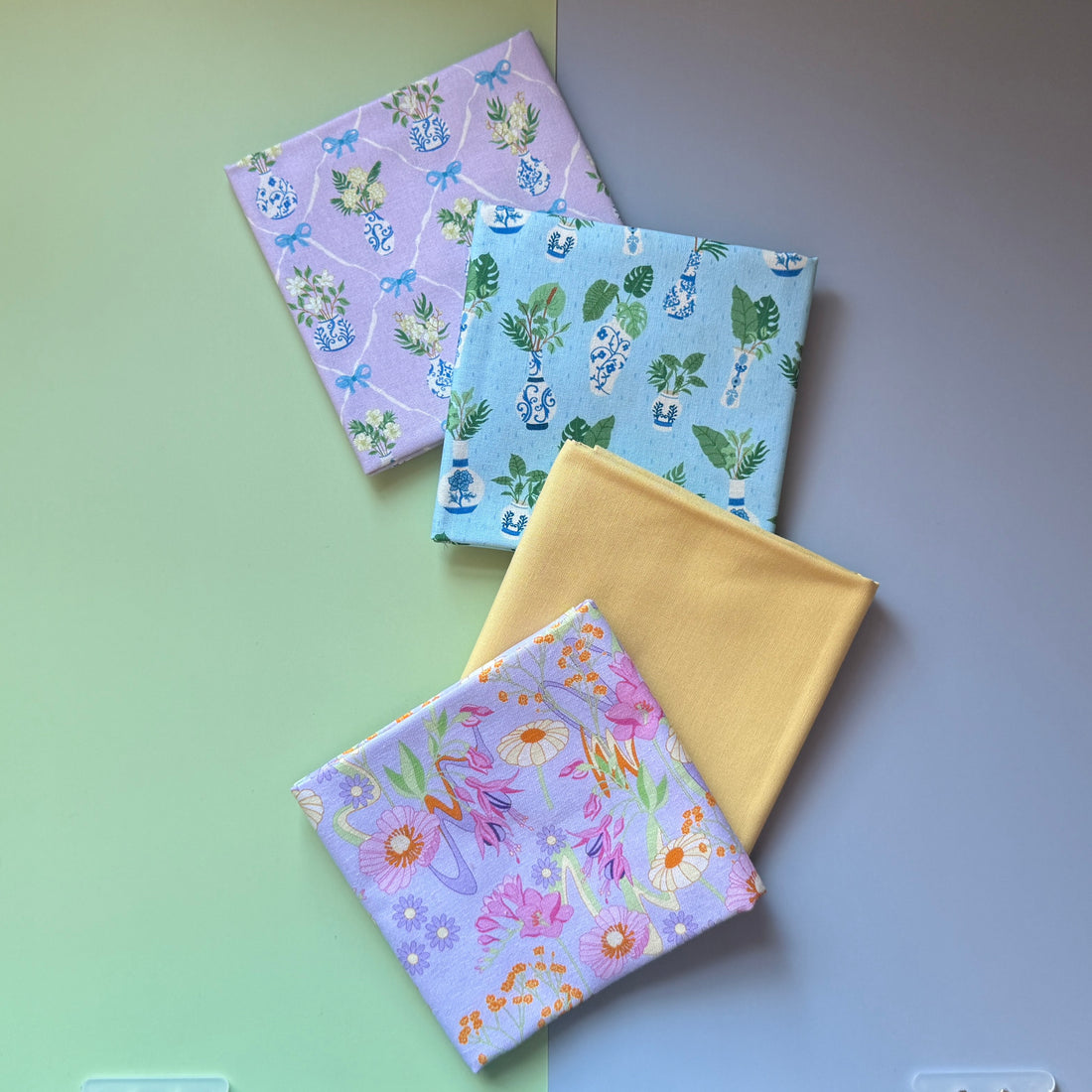 Four folded fat quarters overlapping diagonally: retro florals on lilac, plain yellow and potted plant prints on blue and lilac.