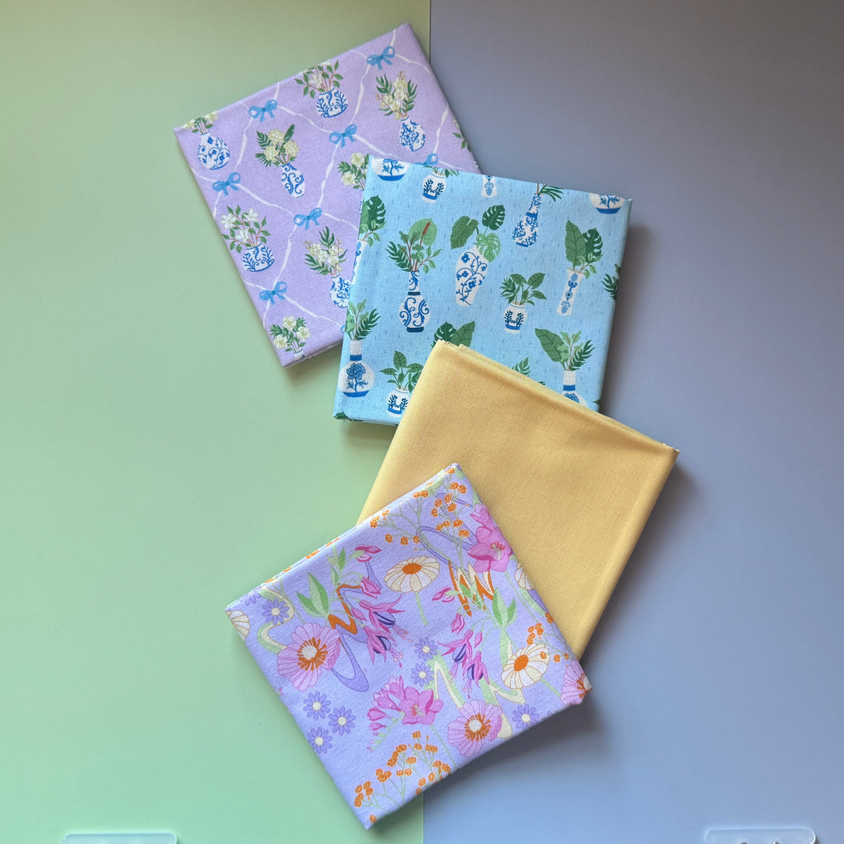 Four folded fat quarters overlapping diagonally: retro florals on lilac, plain yellow and potted plant prints on blue and lilac.
