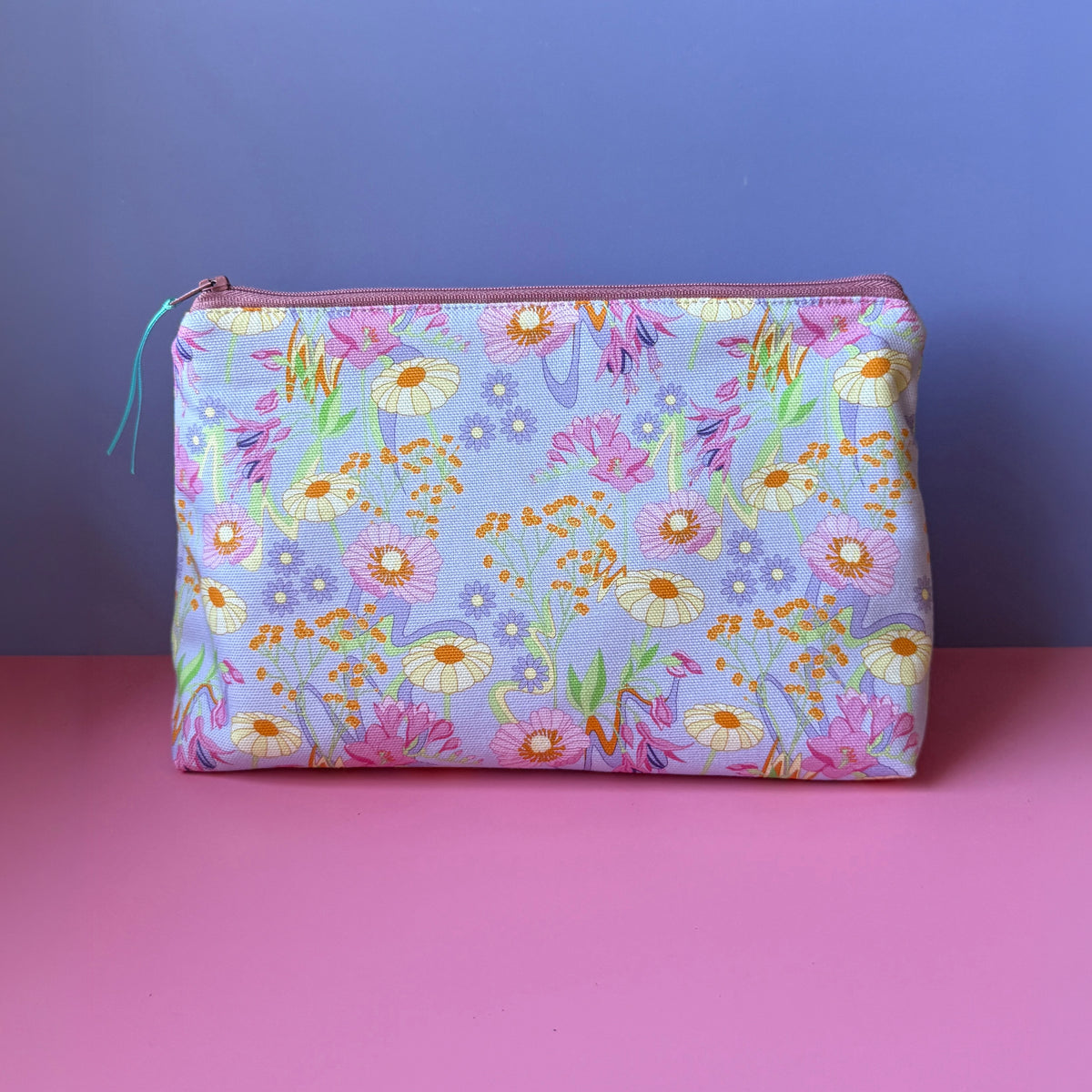 A rectangular zippered pouch with a pastel vintage inspired floral print sits on a pink surface against a lilac background. The pouch features a dense pattern of pink, cream and orange flowers (fuchsias, gypsophelia) on a lilac base, with a  a rose coloured zip. 
