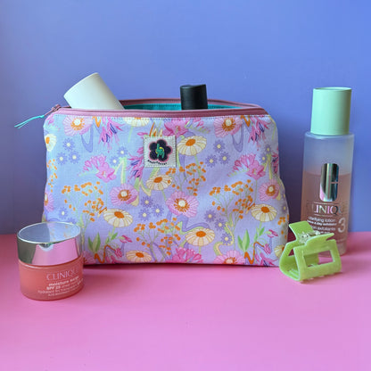 A  zippered wash bag/makeup bag with a pastel vintage inspired floral print sits on a pink surface against a lilac background, surrounded by beauty accessories including a green hair clip, moisturiser bottle and large toner bottle. 